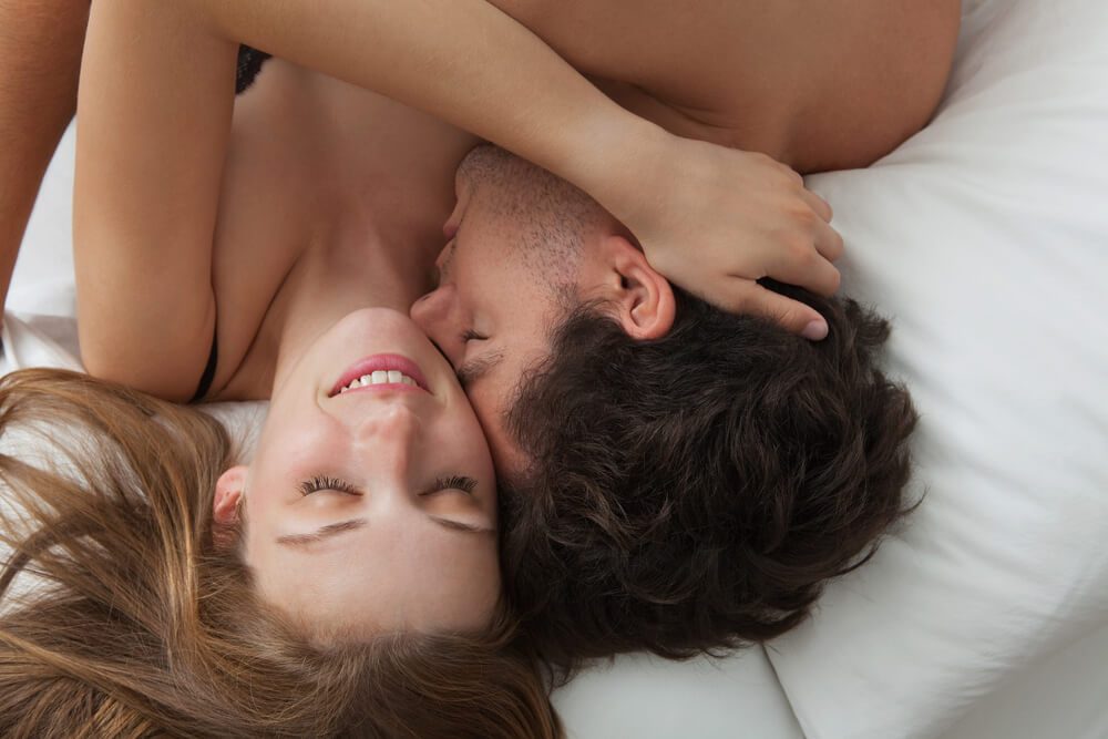 Overhead close up portrait of a young romantic couple hugging and kissing, laying down on a white bed, having sex and loving each other. Love and relationships lifestyle, interior bedroom.