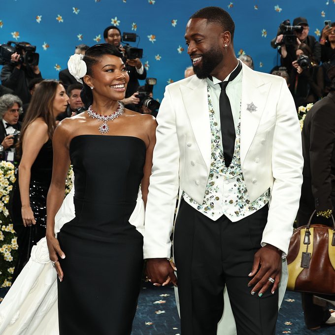 A smiling couple holds hands on a red carpet. The woman wears a strapless black gown and sparkling jewelry, while the man wears a white jacket with a floral vest and black pants. Photographers and a blue, starry backdrop are behind them.
